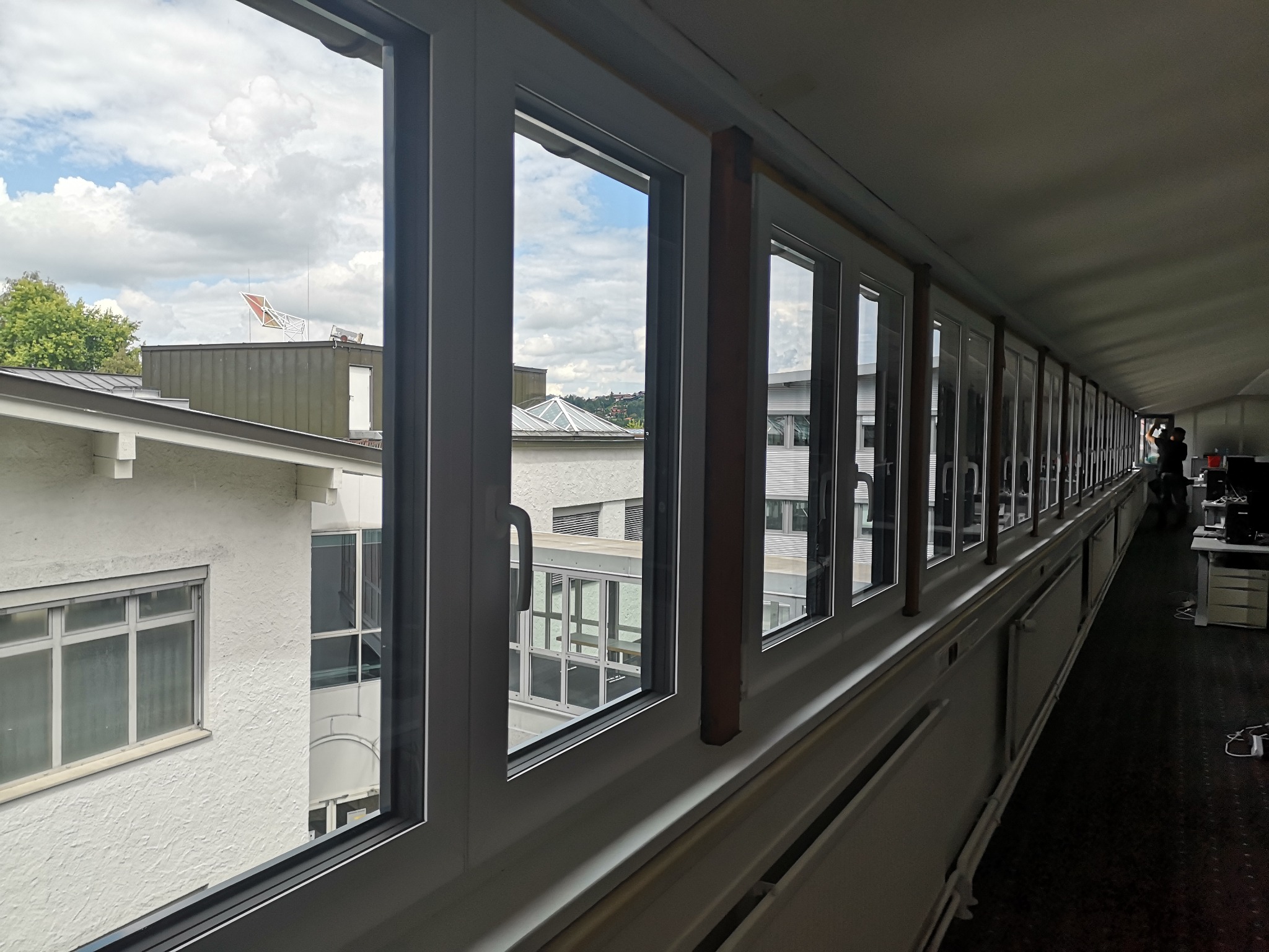 Długi rząd białych okien z widokiem na budynki i niebo z chmurami z wnętrza budynku, widoczny fragment biura.