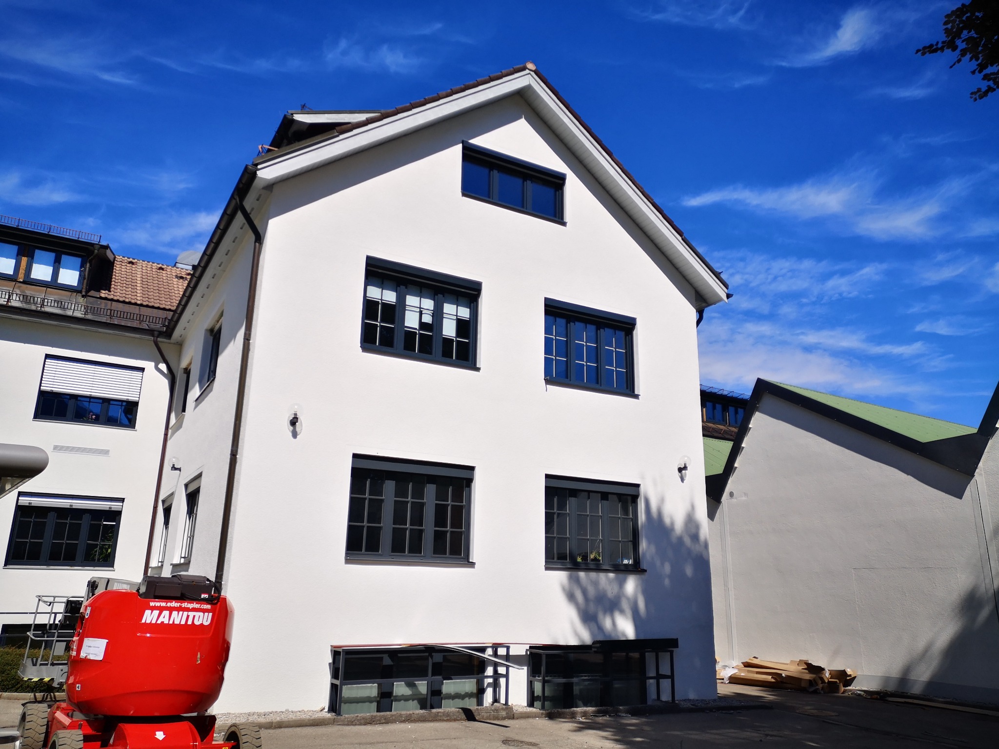 Biały, trzypiętrowy budynek z ciemnymi ramami okiennymi widziany z dołu, z podnośnikiem koszowym Manitou i stertą kartonów w pobliżu.