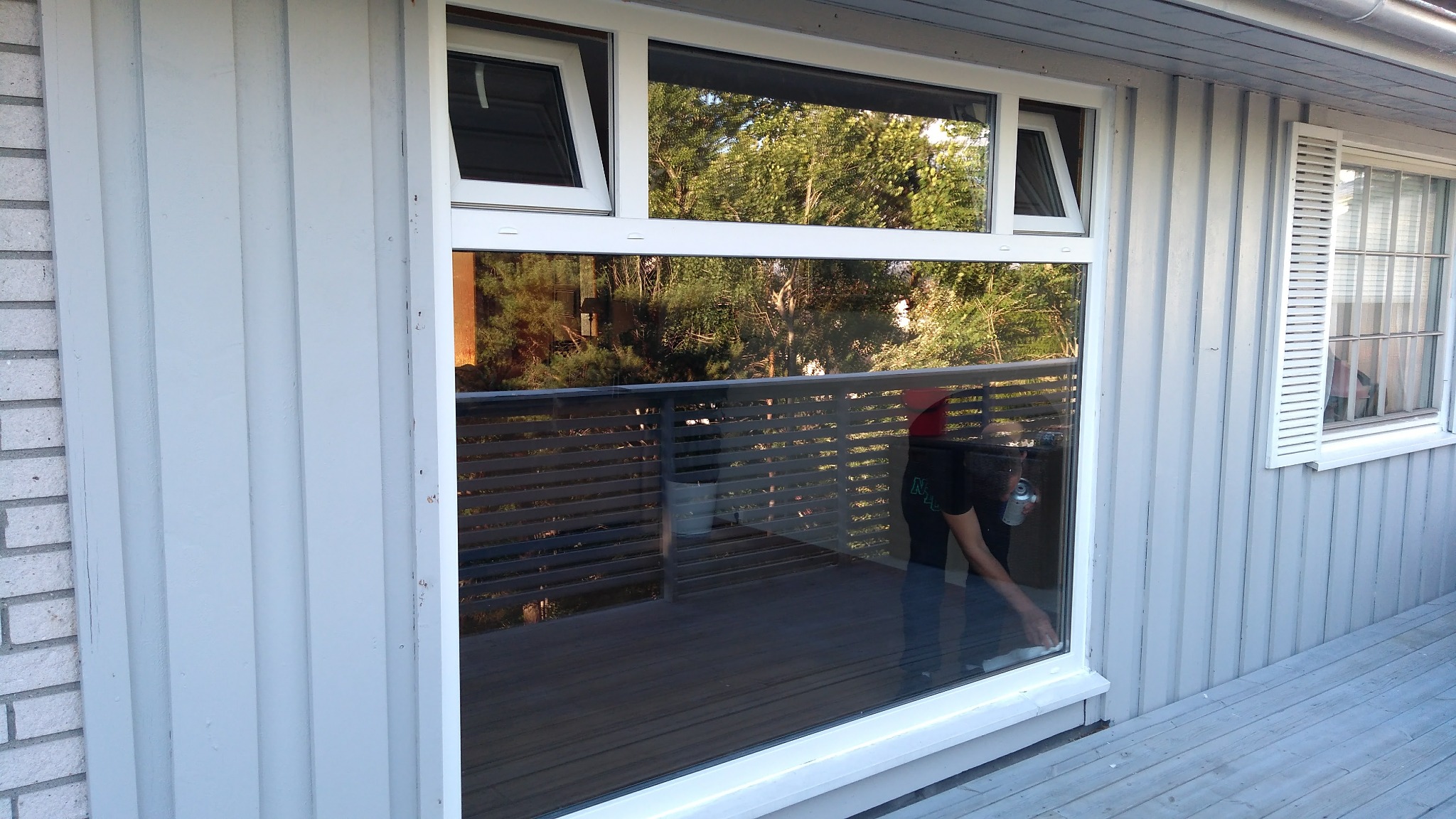 Białe okna PCV z widocznym odbiciem balkonu i zieleni, jedno okno z żaluzjami, ściana zewnętrzna z jasnoszarej deski.