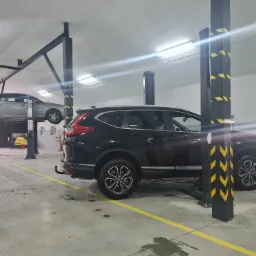 Czarne auto SUV na podnośniku samochodowym w warsztacie, w tle szare auto na drugim podnośniku, żółte linie na podłodze.