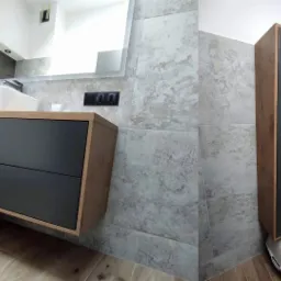 Nowoczesna łazienka z szarą armaturą, wisząca szafka pod umywalką z szufladami w kolorze grafitowym, wysoka szafka łazienkowa w odcieniach drewna, oraz szare płytki imitujące kamień na ścianach...