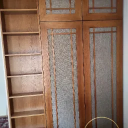 Drewniana szafa z półkami i drzwiami z szarą tkaniną w geometryczne wzory, oświetlona naturalnym światłem. Widoczne logo firmy na dole.