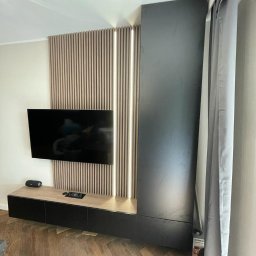 Zacne Meble Piotr Chudawski - Nowoczesny salon z zabudową telewizyjną: pionowe lamele z podświetleniem LED, szafki wiszące z blatem imitującym drewno, wysoka szafa w kolorze grafitowym, parkiet w jodełkę.