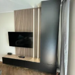 Nowoczesny salon z zabudową telewizyjną: pionowe lamele z podświetleniem LED, szafki wiszące z blatem imitującym drewno, wysoka szafa w kolorze grafitowym, parkiet w jodełkę.