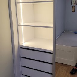 Zacne Meble Piotr Chudawski - Wnętrze białej szafy wnękowej z oświetleniem LED, widoczne półki i szuflady, z boku fragment łóżka i ściany w pokoju.