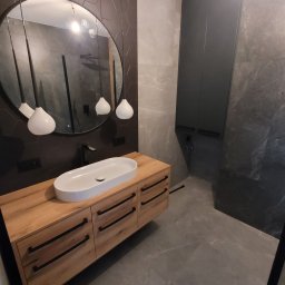Zacne Meble Piotr Chudawski - Nowoczesna łazienka z drewnianą szafką pod umywalkę, owalną białą umywalką nablatową, okrągłym lustrem z trzema wiszącymi lampami, ciemną mozaiką heksagonalną na ścianie i szarą podłogą z płytek...