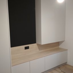 Zacne Meble Piotr Chudawski - Nowoczesna zabudowa ściany z szafkami i blatem z jasnego drewna oraz czarnym panelem ściennym z gniazdkami elektrycznymi.