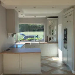 Jasna kuchnia z wyspą i widokiem na zieleń za oknem; drewniany blat, białe fronty szafek, dekoracyjna podłoga, surowa żarówka zwisająca z sufitu.