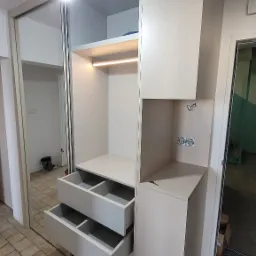 Biała szafa wnękowa z przesuwnymi drzwiami lustrzanymi i otwartymi szufladami, widoczne gniazdka elektryczne na bocznej ściance, oświetlenie LED wewnątrz.