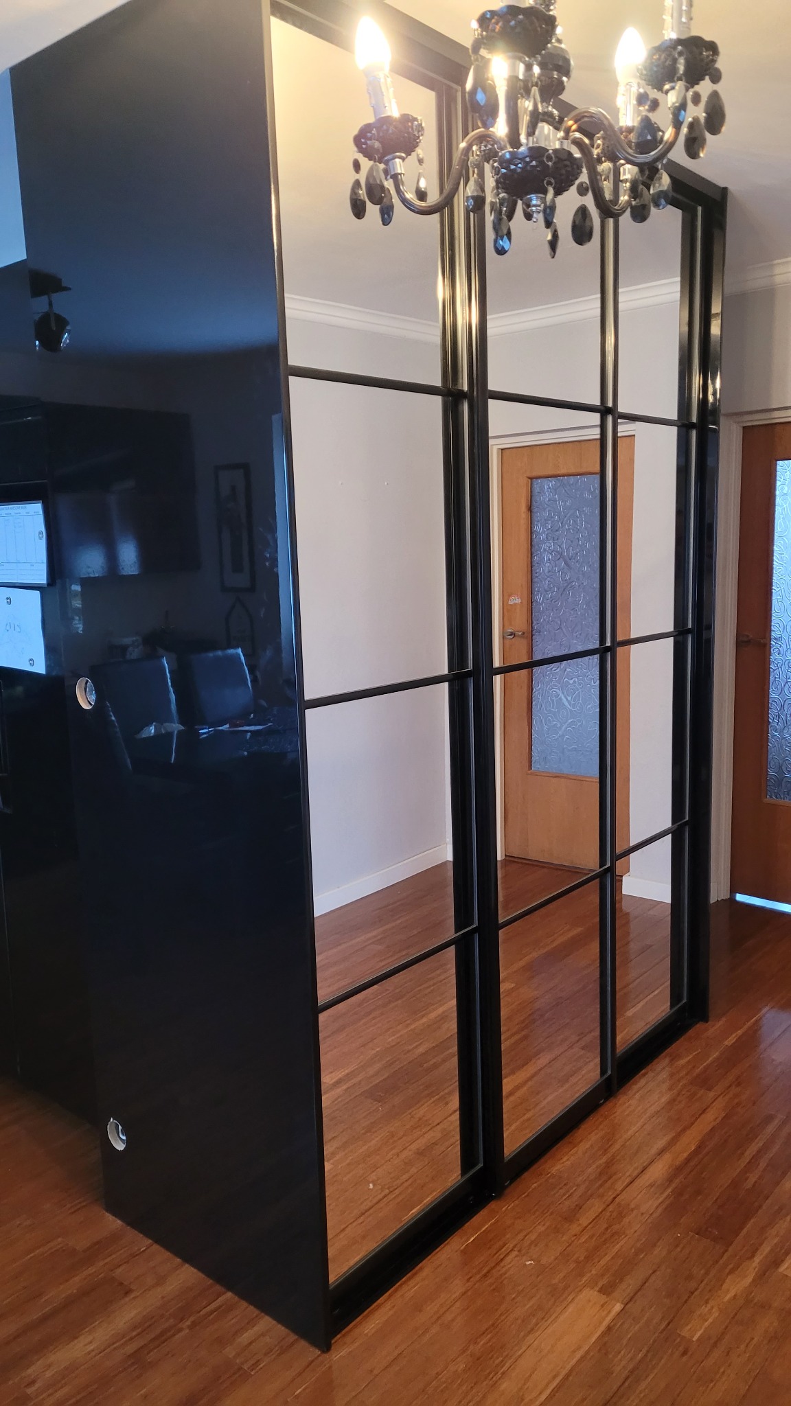 Czarna szafa przesuwna z lustrami i czarnymi szprosami, odbijająca kryształowy żyrandol i wnętrze pokoju z drewnianą podłogą.