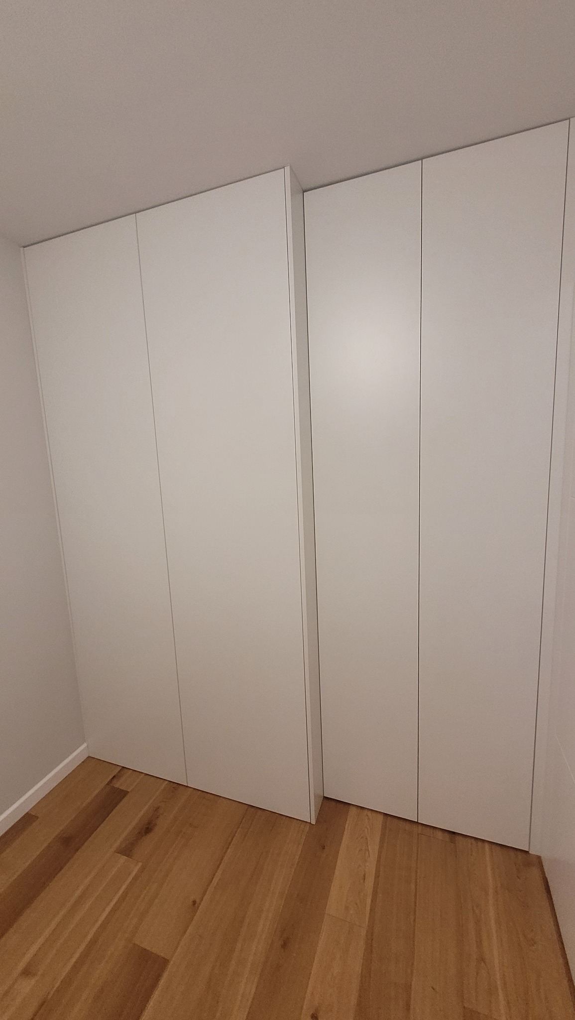 Białe szafy wnękowe z gładkimi frontami, dopasowane do wysokości pomieszczenia, na podłodze z paneli drewnianych.