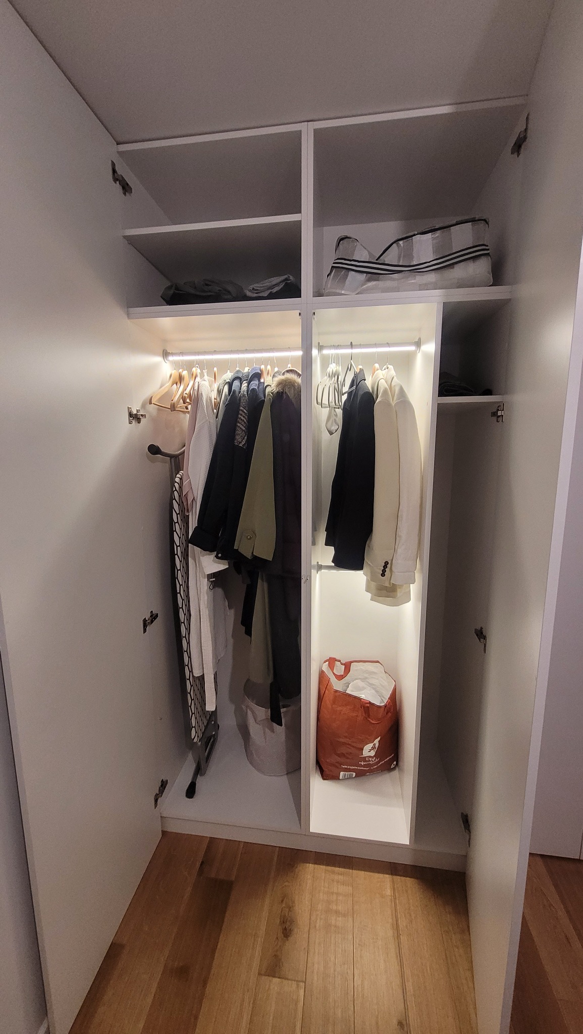 Wnętrze białej szafy z oświetleniem LED, ubrania wiszące na wieszakach, deska do prasowania po lewej stronie, torba na zakupy na dolnej półce po prawej stronie, drewniana podłoga.