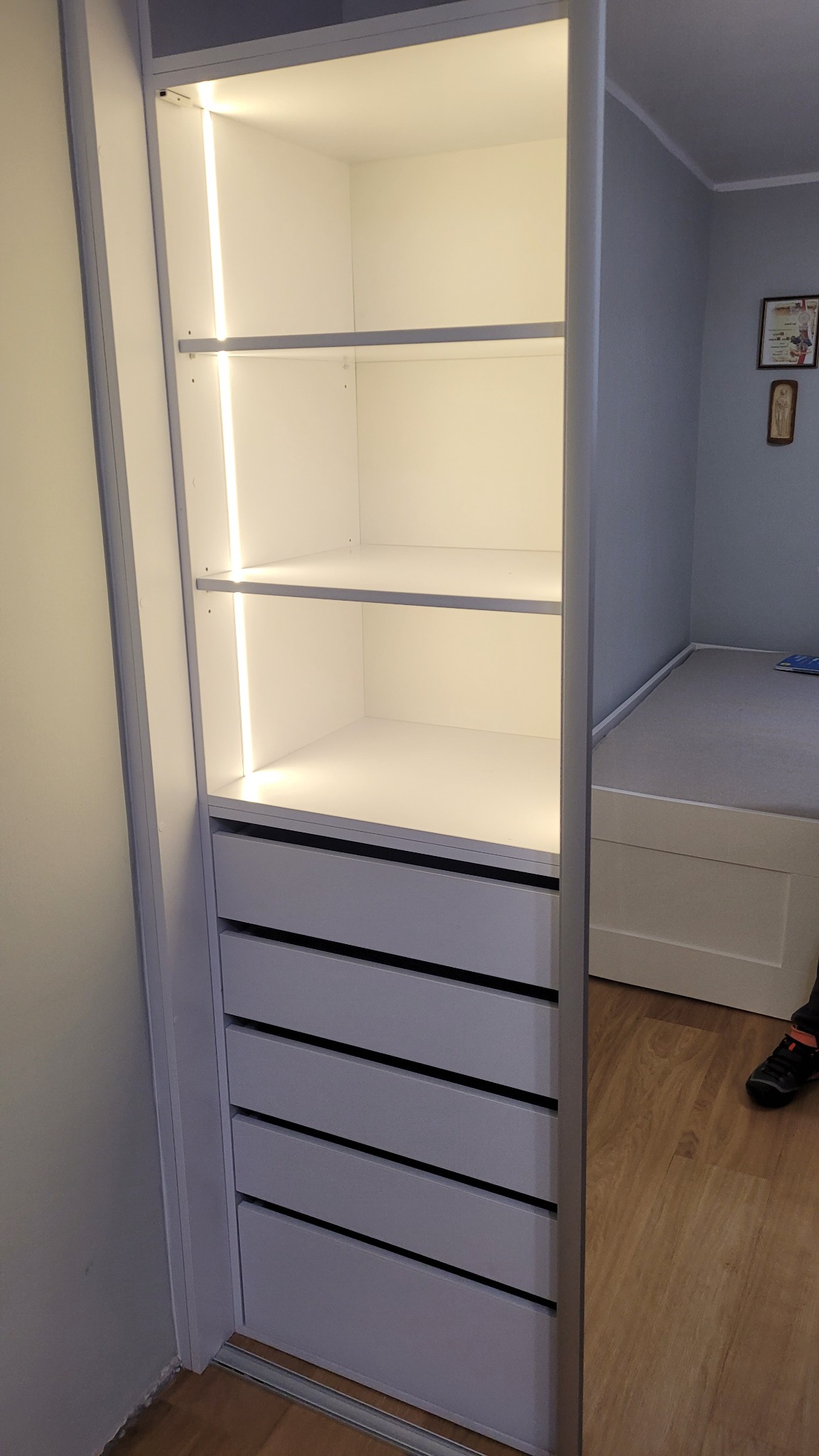 Wnętrze białej szafy wnękowej z oświetleniem LED, widoczne półki i szuflady, z boku fragment łóżka i ściany w pokoju.