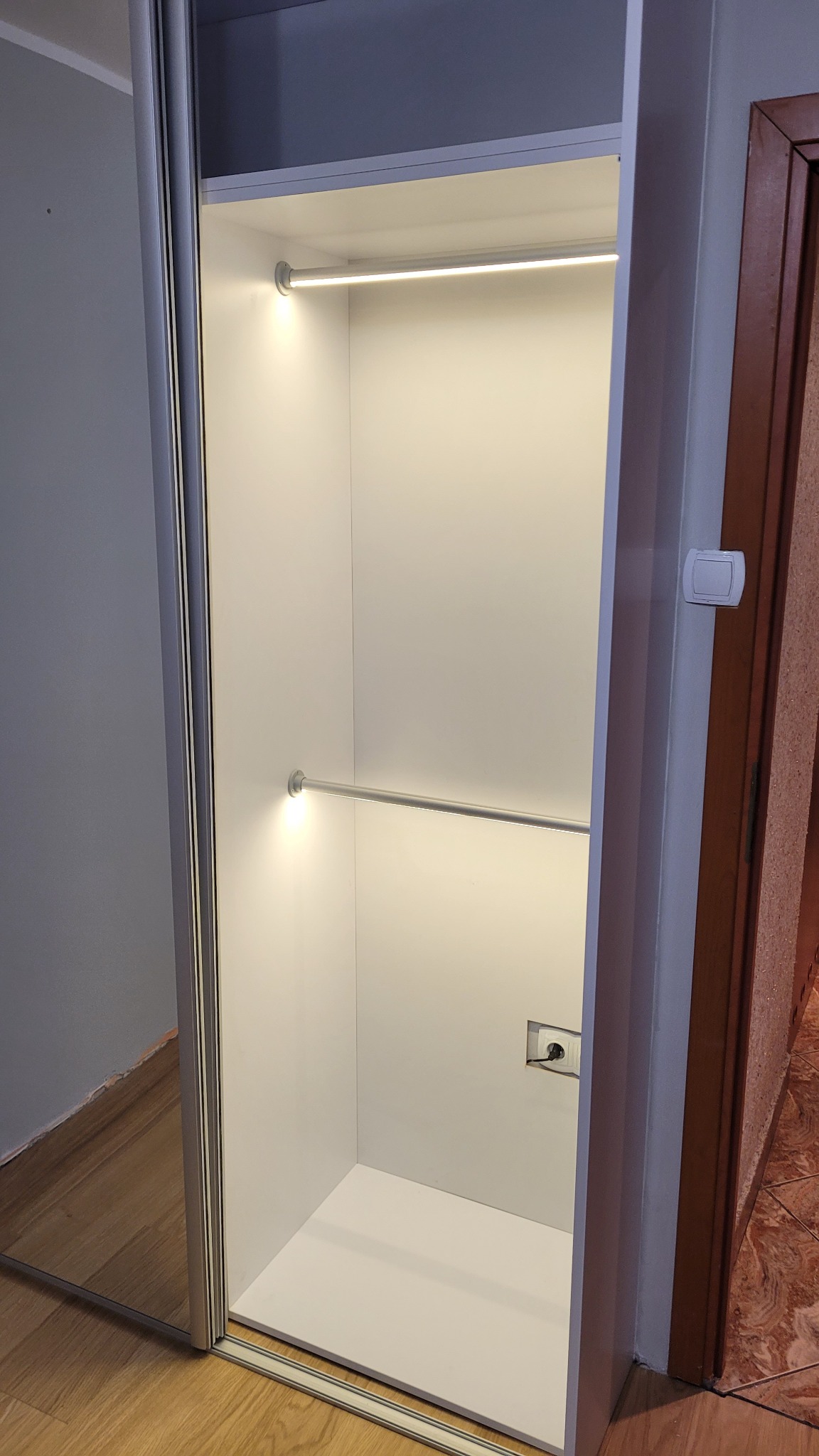 Wnętrze szafy wnękowej z przesuwnymi drzwiami, oświetleniem LED i gniazdkiem elektrycznym, widoczne szyny drzwi.