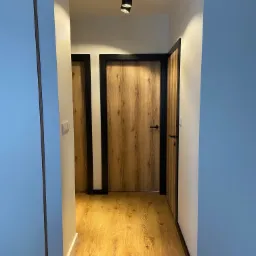 Wnętrze korytarza z drewnianą podłogą i dwiema parami drzwi z czarnymi framugami, oświetlone punktowymi lampami sufitowymi.