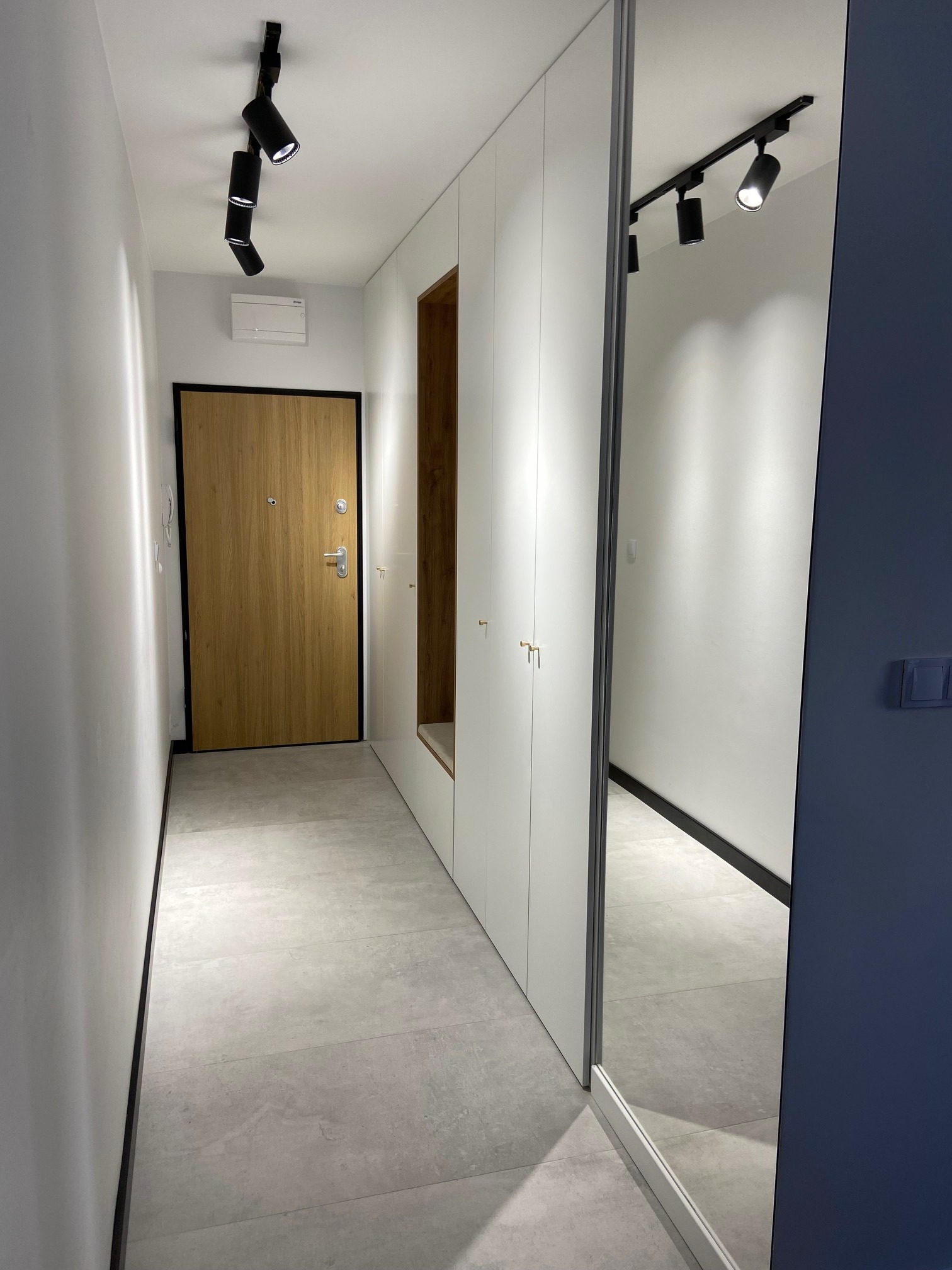 Nowoczesny przedpokój z jasnymi ścianami, szarą podłogą z płytek, drewnianymi drzwiami wejściowymi z czarną ościeżnicą, białymi szafami w zabudowie z wnęką na siedzisko i dużym lustrem.