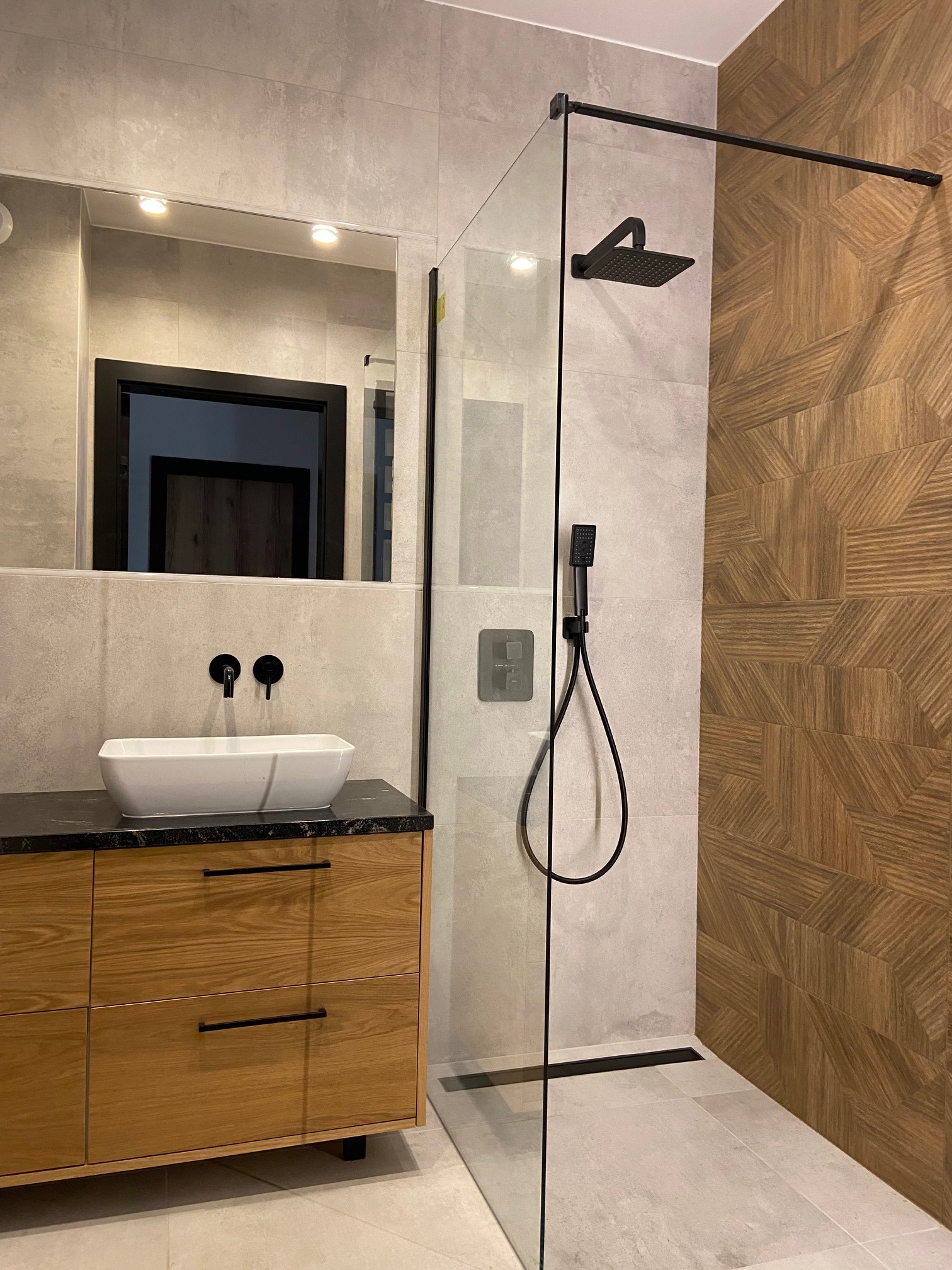 Nowoczesna łazienka z kabiną prysznicową typu walk-in, drewnianą szafką pod umywalką z czarnym blatem, geometryczną okładziną ścienną z drewna i czarną armaturą.