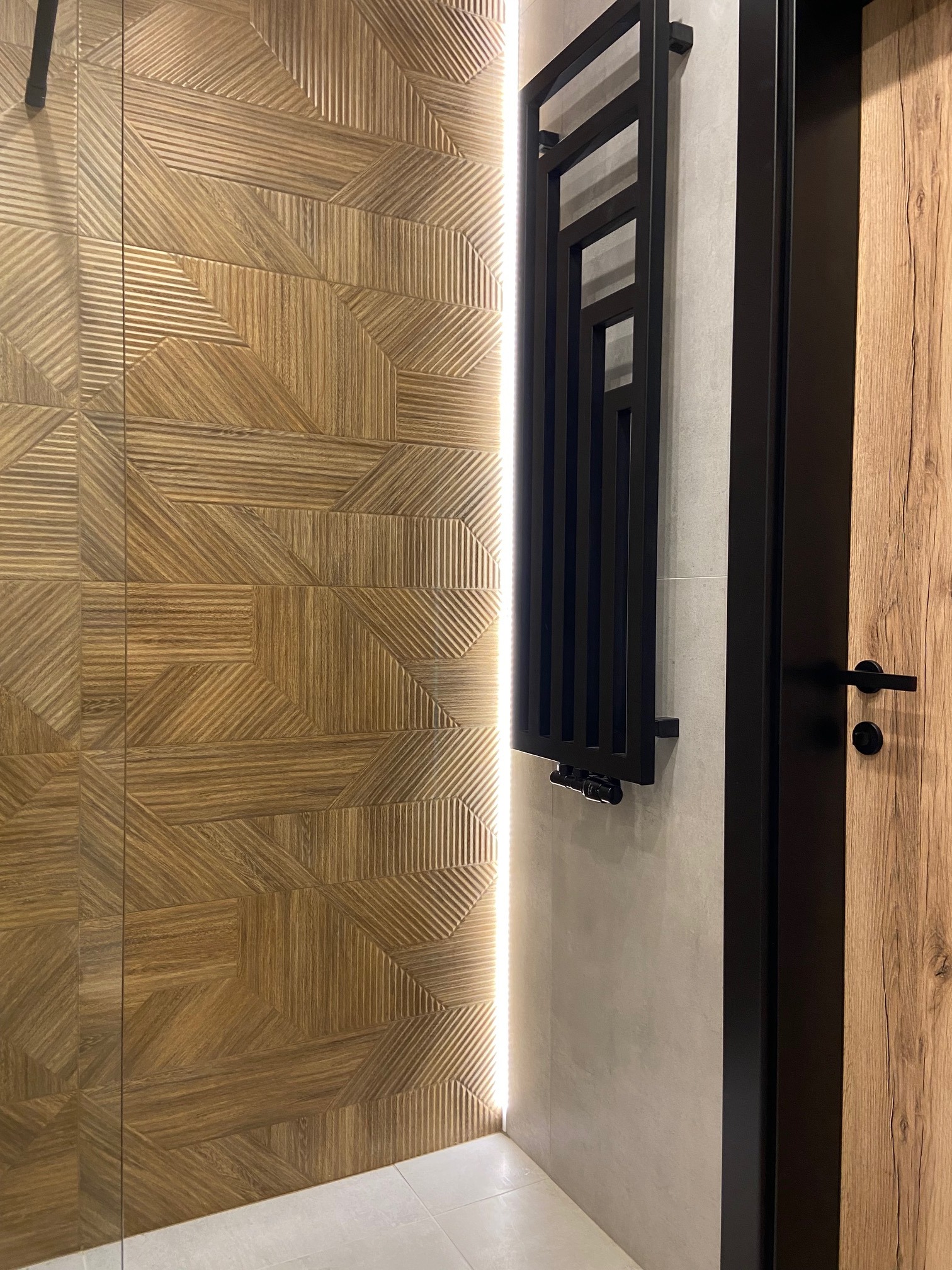 Nowoczesna łazienka z drewnopodobną ścianą z płytek o geometrycznym wzorze, podświetleniem LED, czarnym grzejnikiem drabinkowym i drewnianymi drzwiami z czarną klamką.