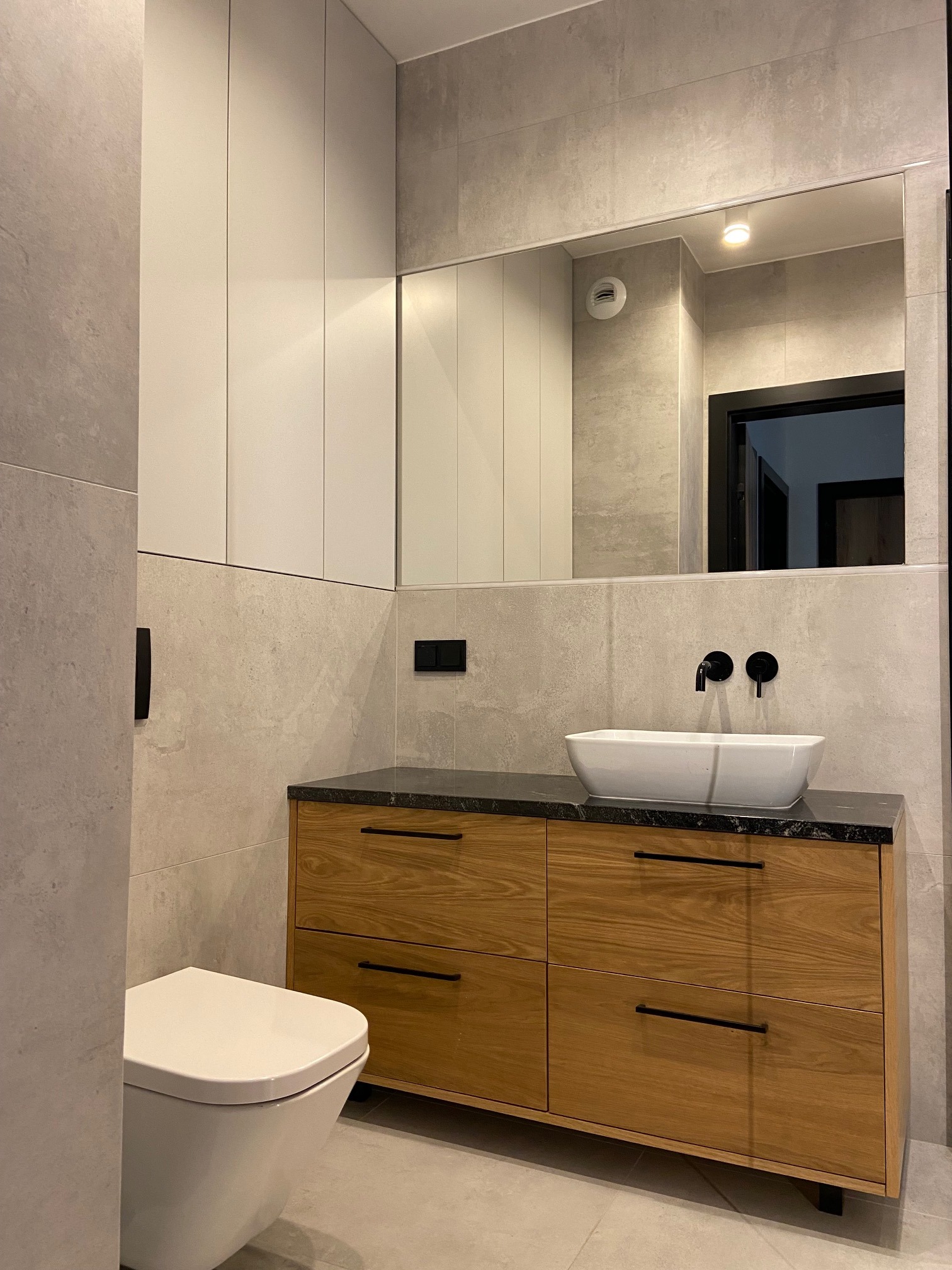 Nowoczesna łazienka z szarą płytką, drewnianą szafką pod umywalką z czarnym blatem, białą umywalką nablatową, czarną armaturą i wiszącą toaletą.