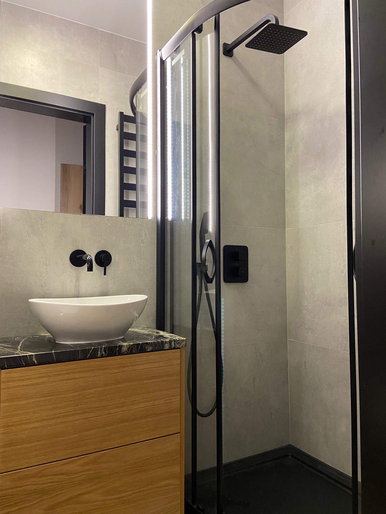 Nowoczesna łazienka z kabiną prysznicową typu walk-in z czarnymi elementami, umywalką nablatową na szafce z drewnianym frontem i blatem z ciemnego marmuru, szare płytki na ścianach i oświetlenie LED.