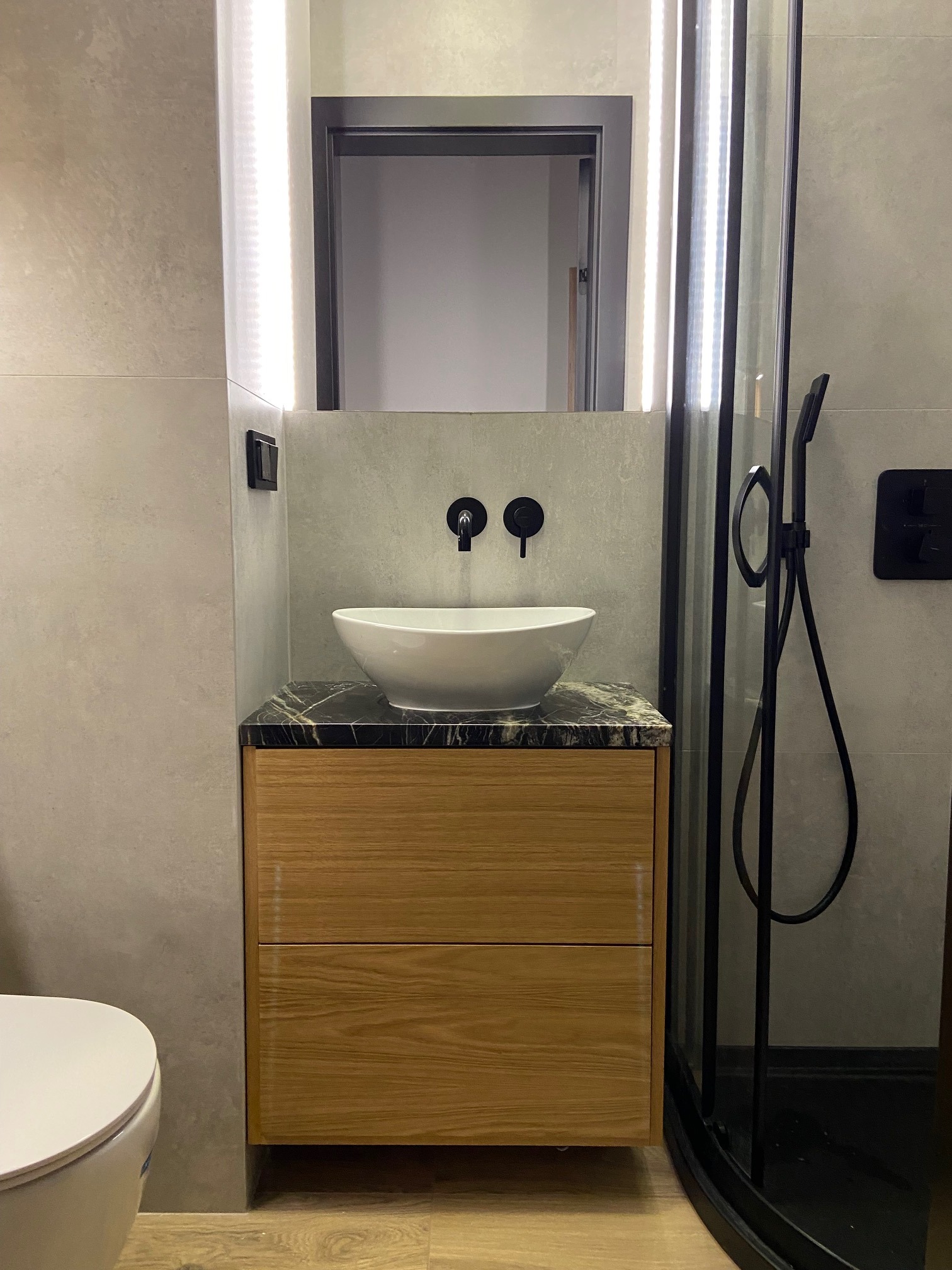 Nowoczesna łazienka z umywalką nablatową na szafce z drewnianymi szufladami, czarną armaturą i kabiną prysznicową z czarnymi profilami, oświetlenie LED wokół lustra.