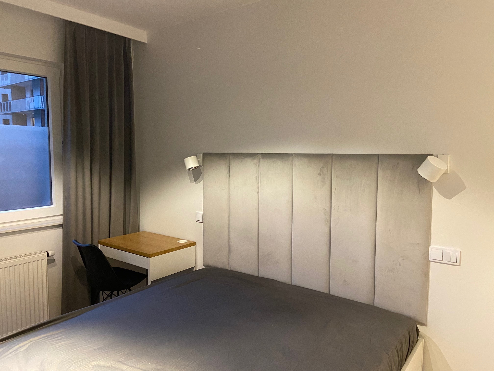 Nowoczesna sypialnia z szarym łóżkiem, tapicerowanym wezgłowiem, biurkiem z drewnianym blatem i szarą zasłoną przy oknie z widokiem na budynek.