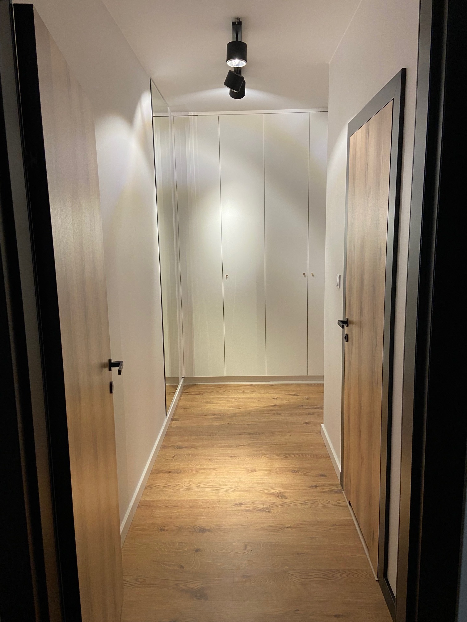 Wnętrze korytarza z drewnianą podłogą, białymi szafami w zabudowie i drzwiami z okleiną drewnopodobną w czarnych ramach, oświetlenie punktowe na suficie.