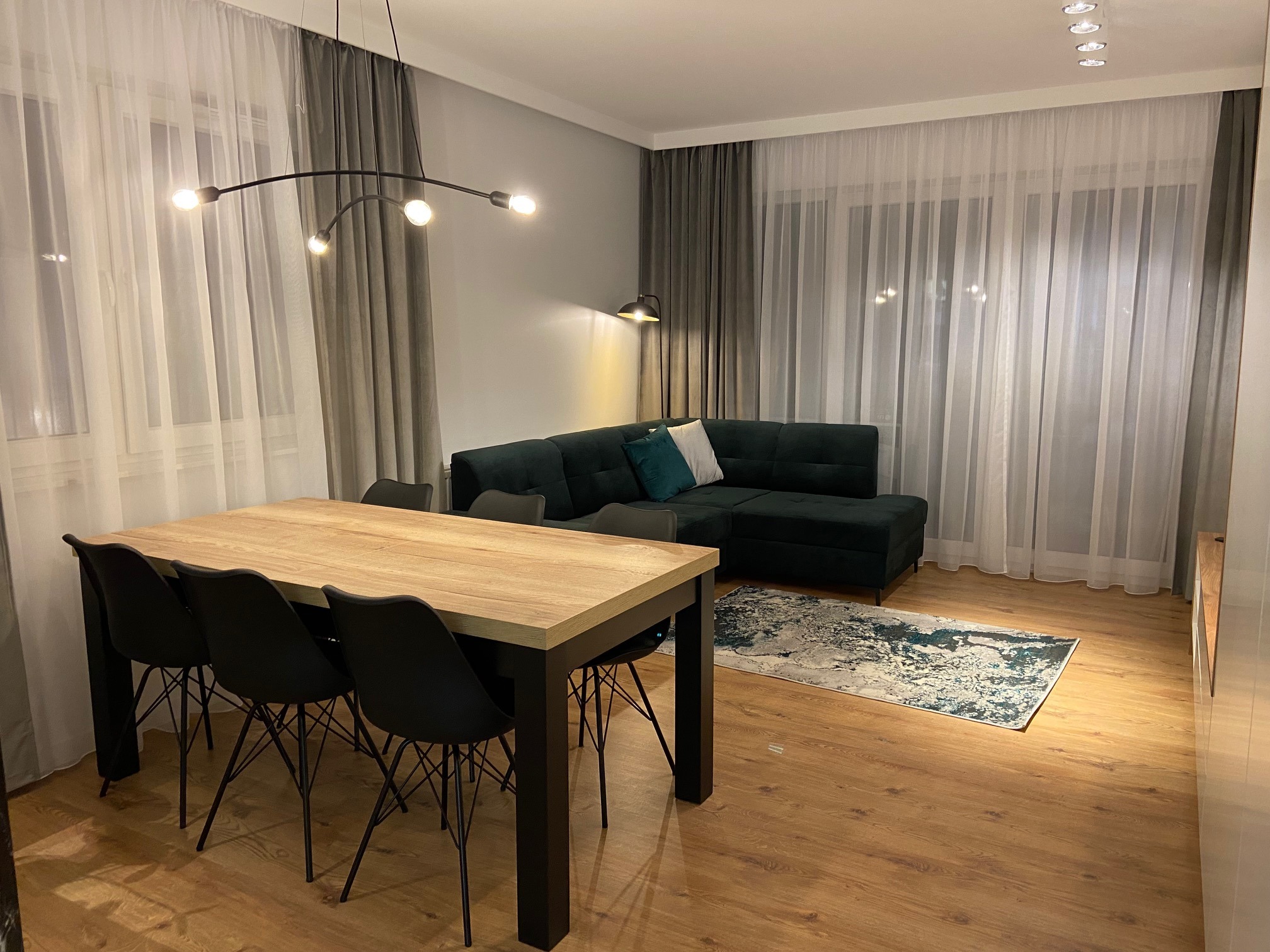 Jasne wnętrze salonu z drewnianą podłogą, dużym stołem z czarnymi krzesłami, ciemnozieloną sofą narożną, białymi i szarymi zasłonami oraz nowoczesną lampą sufitową.
