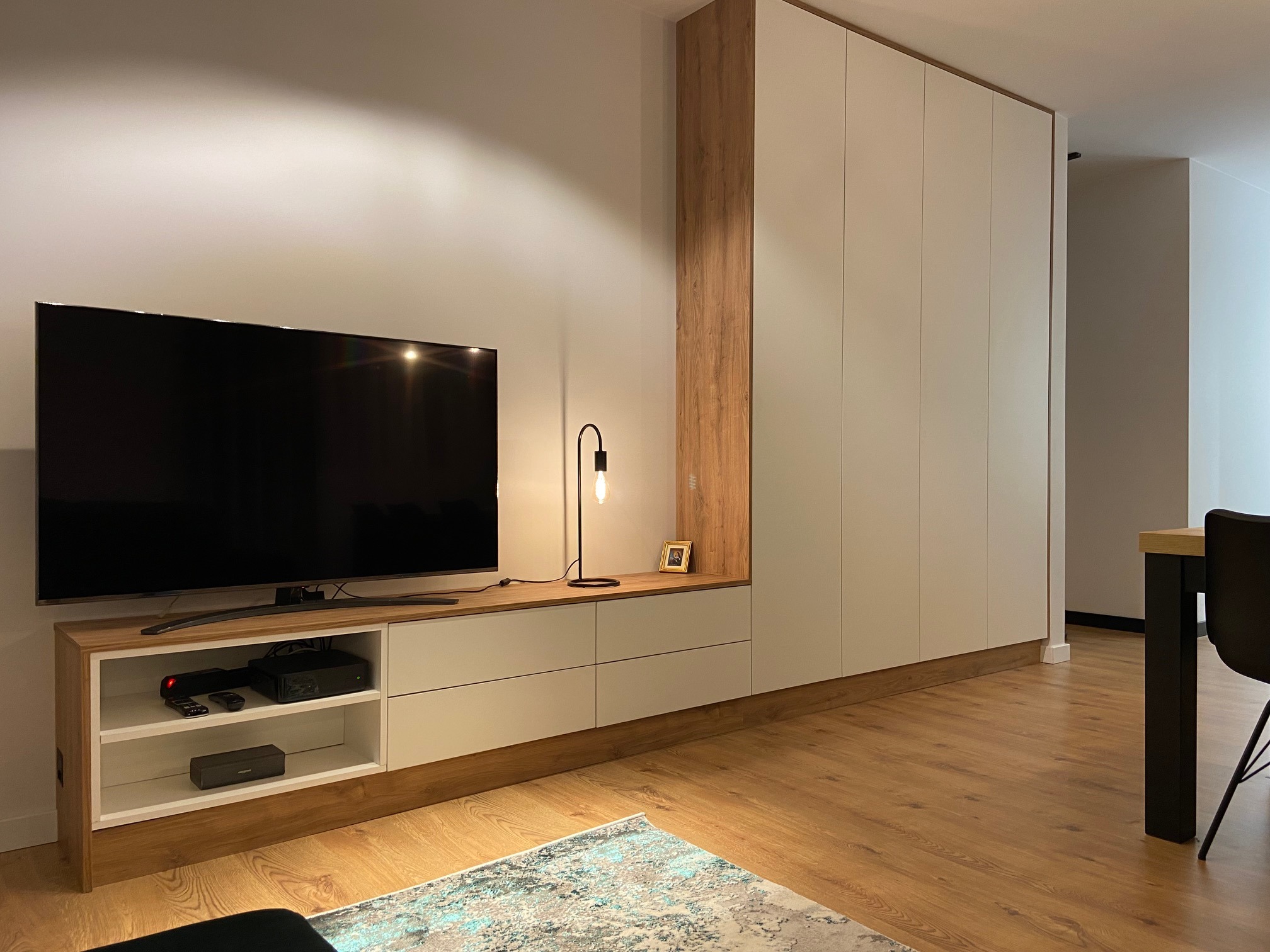 Nowoczesny salon z zabudową meblową: białe szafy z drewnianymi akcentami, telewizor na szafce RTV z otwartymi półkami, lampka biurkowa i dywan w odcieniach szarości i błękitu na drewnianej podłodze.