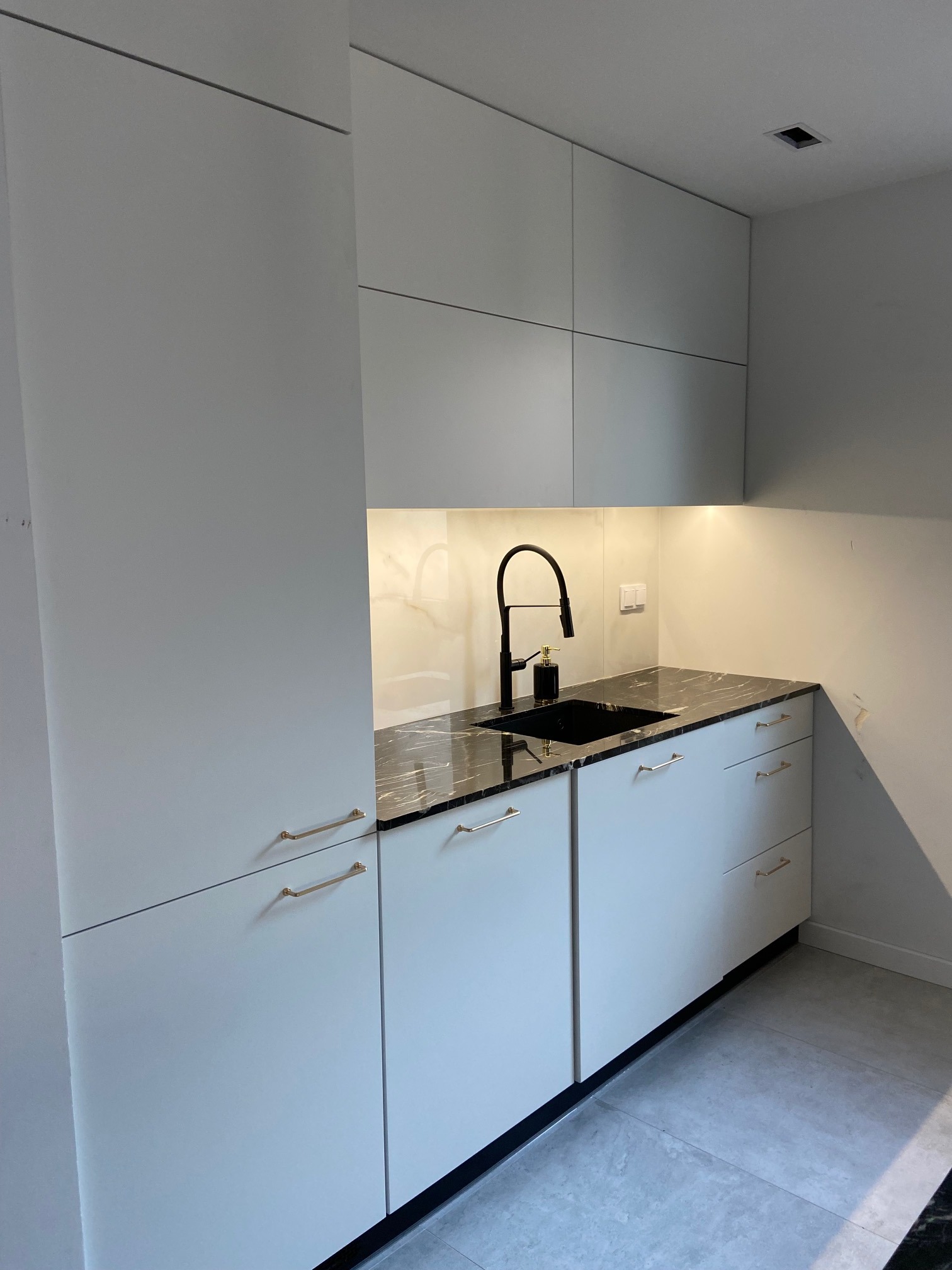 Nowoczesna kuchnia z białymi szafkami, czarnym blatem imitującym marmur i złotymi uchwytami, czarna armatura, oświetlenie punktowe.