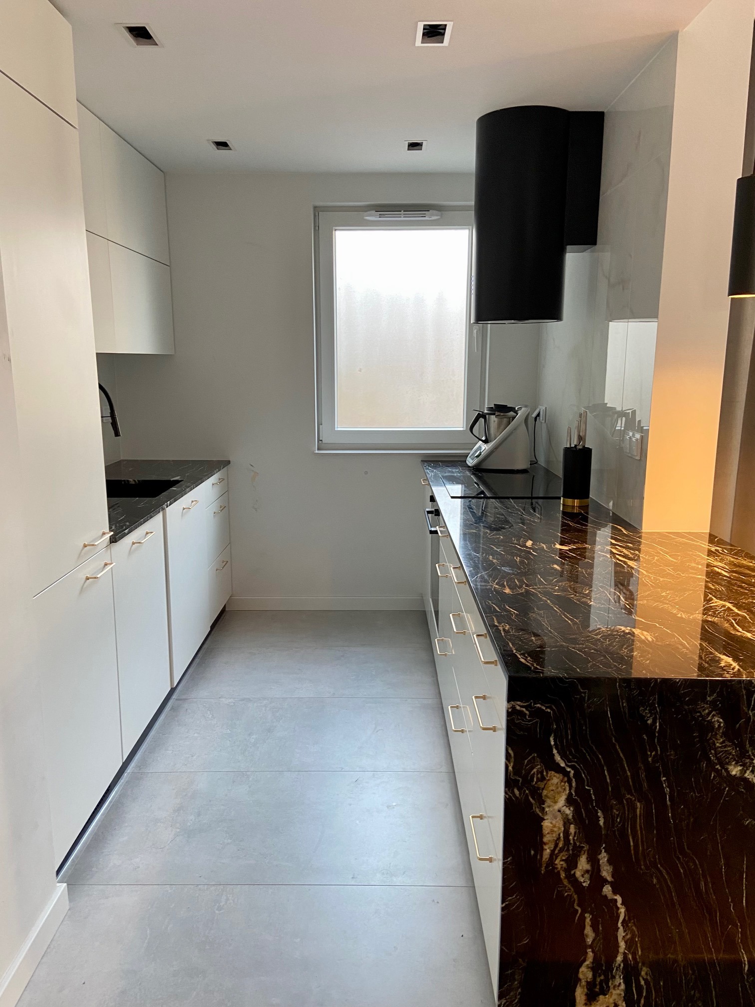 Nowoczesna kuchnia z białymi szafkami, czarnym blatem z marmurowym wzorem, czarnym okapem i szarymi płytkami podłogowymi, oświetlona światłem dziennym wpadającym przez okno.