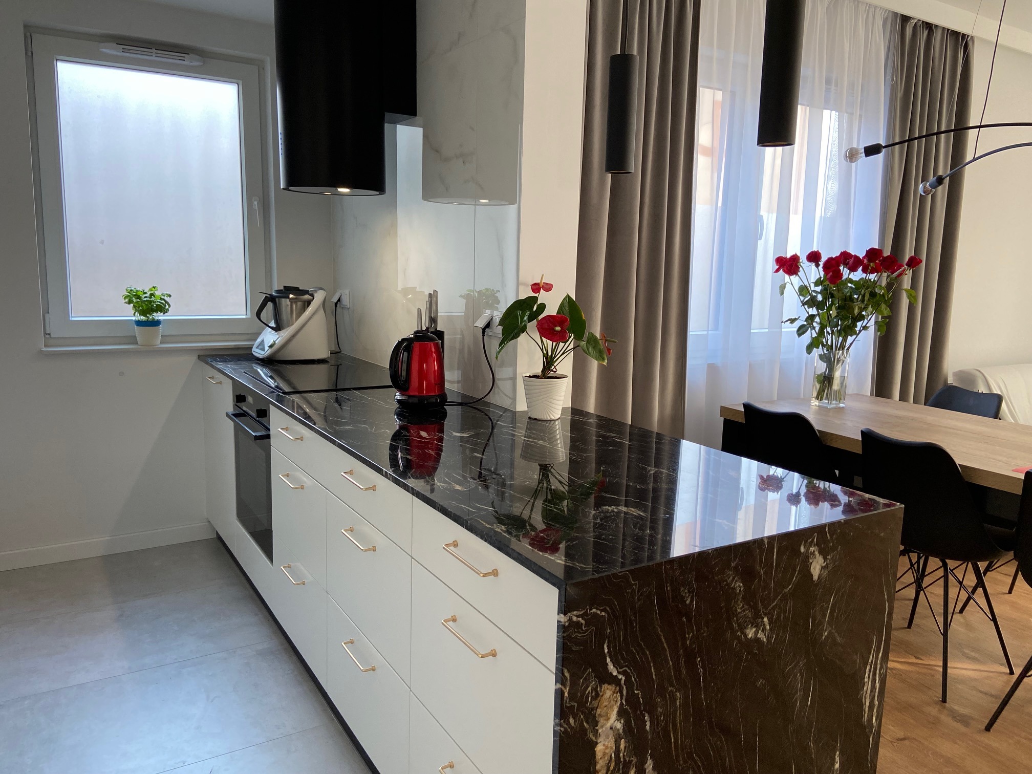 Nowoczesna kuchnia z czarnym marmurowym blatem, białymi szafkami z złotymi uchwytami, czerwonym czajnikiem i kwiatami w wazonie na stole w jadalni w tle.