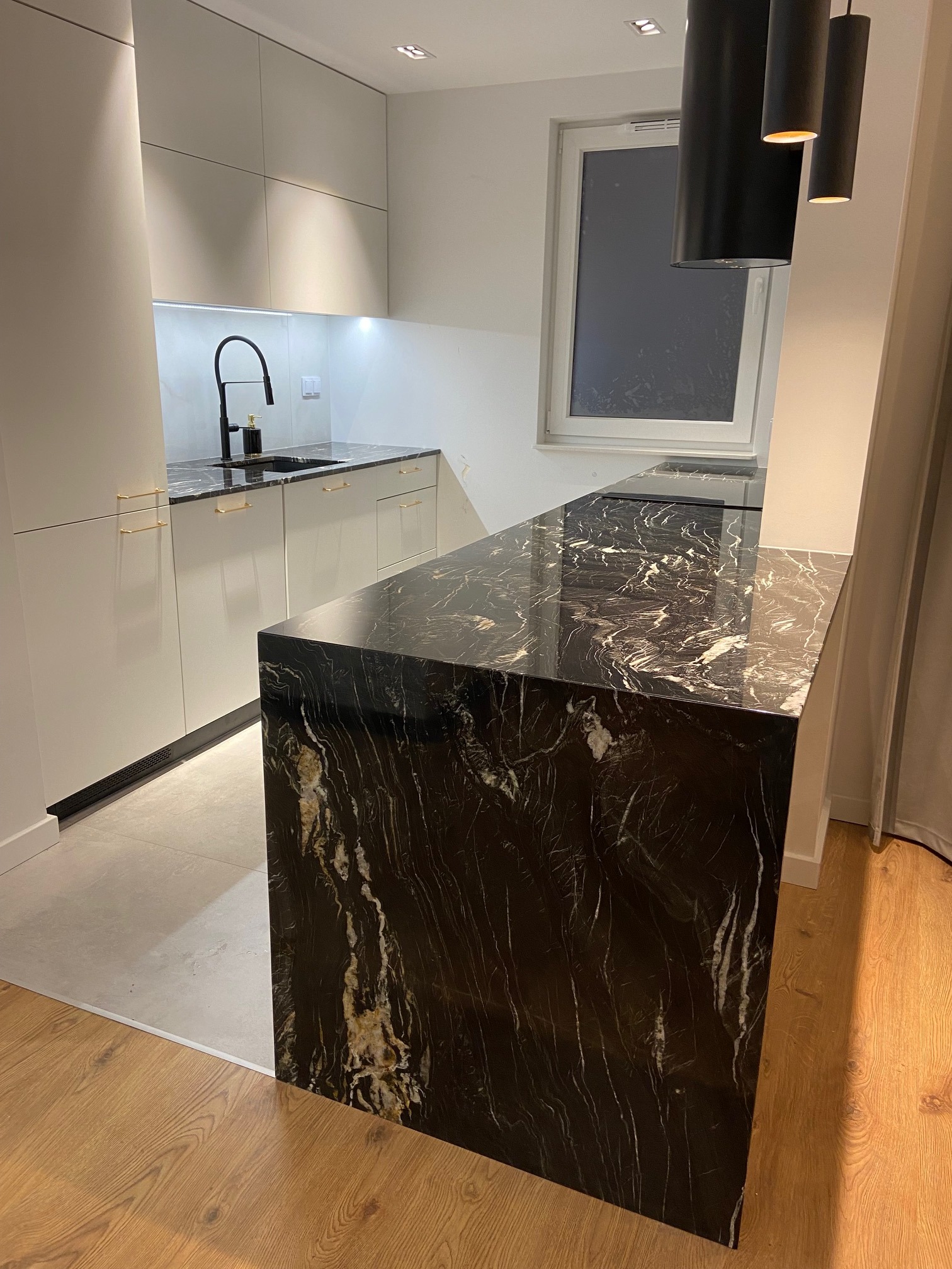 Nowoczesna kuchnia z białymi szafkami, czarnym blatem z marmuru i czarną armaturą, oświetlona trzema wiszącymi lampami.