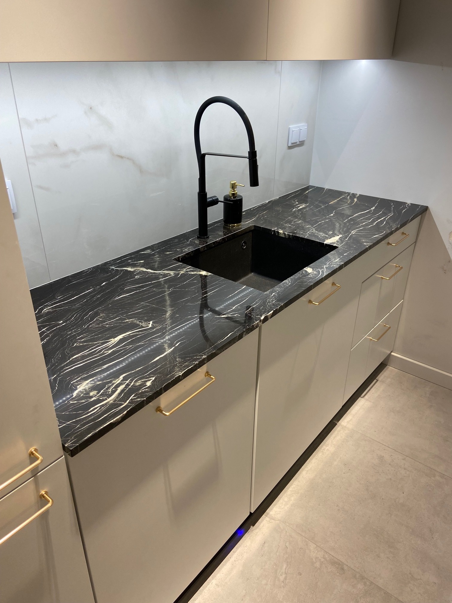 Nowoczesna kuchnia z czarnym marmurowym blatem i złotymi uchwytami, minimalistyczna armatura, białe fronty szafek i ceramiczna ściana.