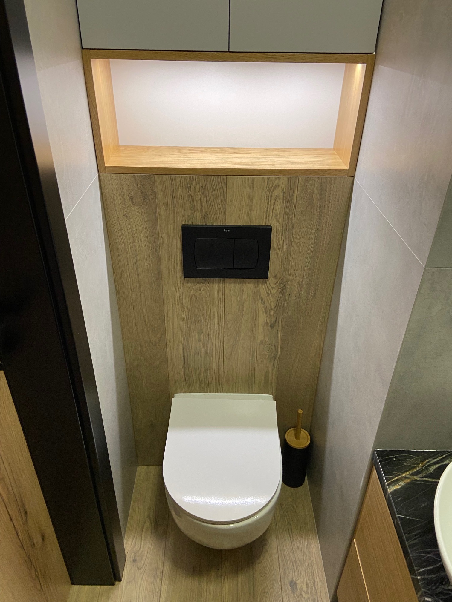 Nowoczesna toaleta w odcieniach szarości i drewna, z podświetlaną wnęką nad spłuczką i czarnym przyciskiem Roca, widoczna szczotka do WC i fragment blatu umywalki z czarnego marmuru.
