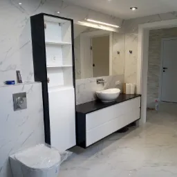 Nowoczesna łazienka z białymi marmurowymi płytkami, wiszącą szafką z umywalką nablatową, toaletą w folii ochronnej i wysoką szafką z czarnymi bokami.