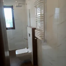 Nowoczesna łazienka z wiszącą toaletą, kabiną prysznicową z deszczownicą i białym grzejnikiem drabinkowym na ścianie z marmurowymi płytkami.