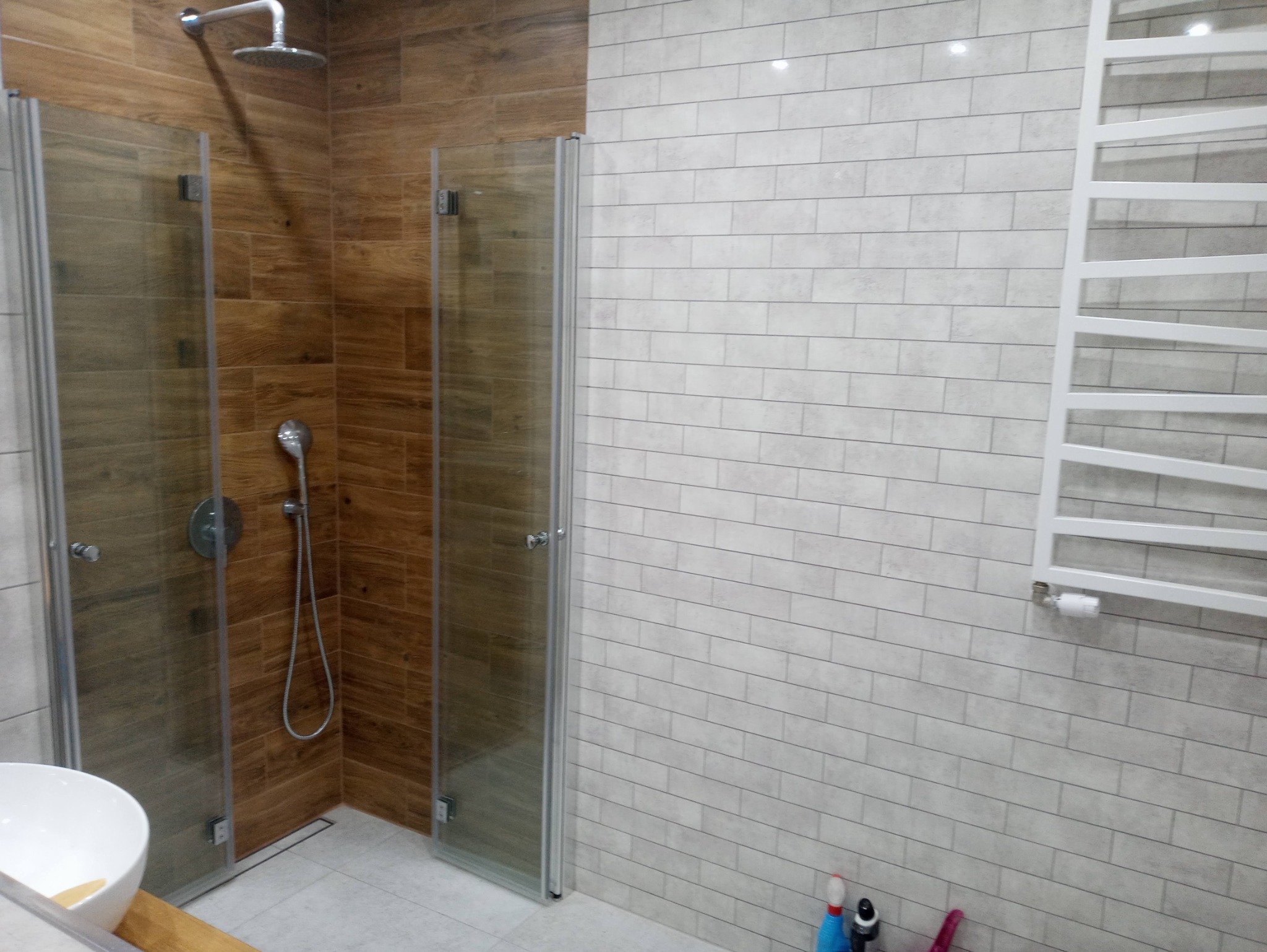 Nowoczesna kabina prysznicowa z deszczownicą i ścianami wyłożonymi płytkami imitującymi drewno oraz białymi płytkami cegiełkami, z widocznym grzejnikiem drabinkowym i fragmentem umywalki.