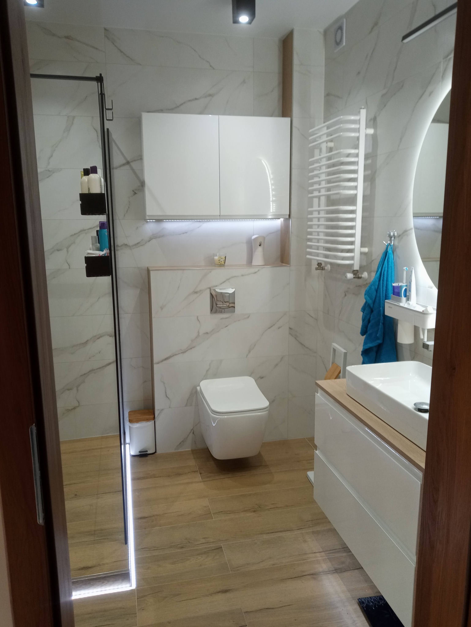 Nowoczesna łazienka z wiszącą miską WC, szafką nad nią z podświetleniem LED, grzejnikiem drabinkowym i umywalką nablatową na białej szafce z drewnianym blatem, wykończona płytkami imitującymi...