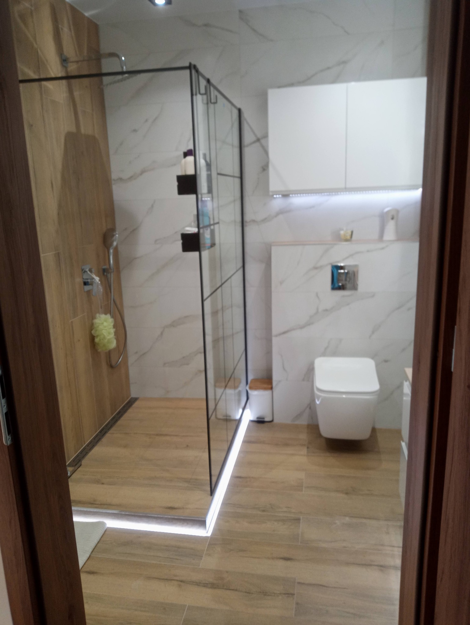 Nowoczesna łazienka z kabiną prysznicową typu walk-in z czarnymi profilami, płytkami imitującymi drewno na podłodze i ścianie prysznica, oraz białymi płytkami imitującymi marmur na pozostałych...