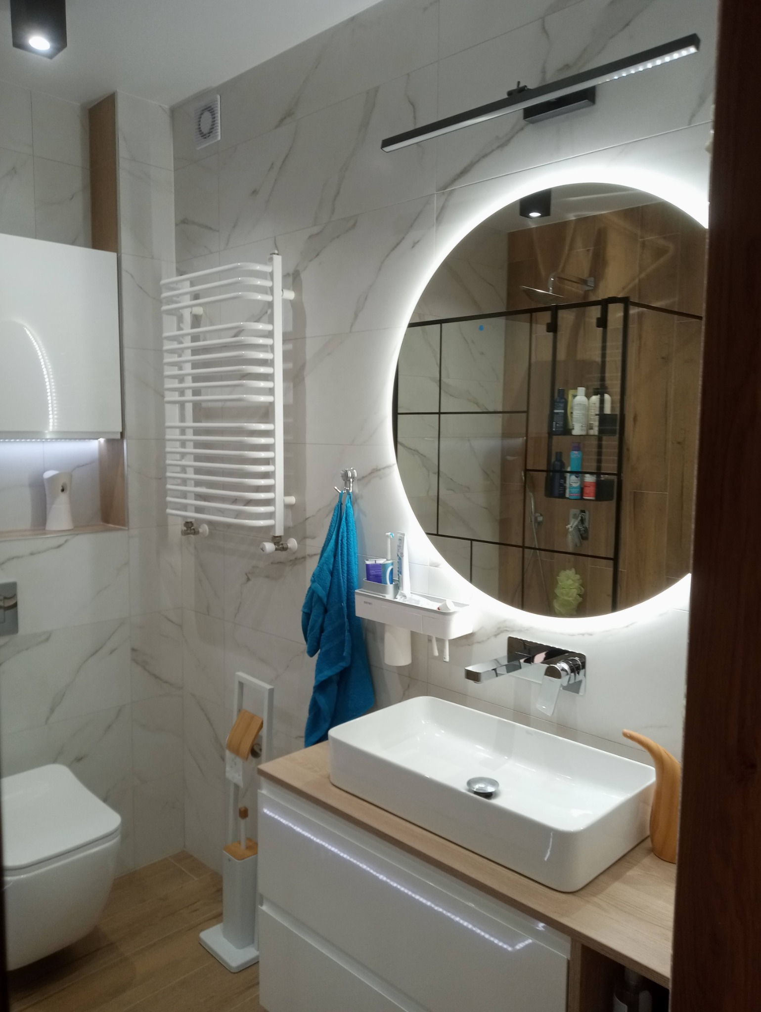 Nowoczesna łazienka z białymi marmurkowymi płytkami, podświetlanym okrągłym lustrem, prostokątną umywalką nablatową i grzejnikiem drabinkowym.