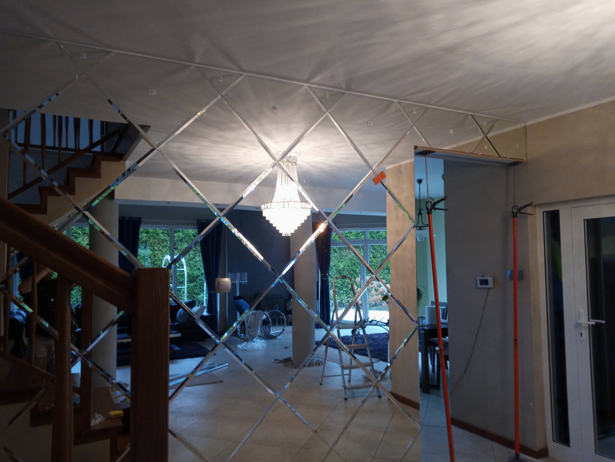 Montaż lustrzanej ściany z romboidalnych elementów w przedpokoju, widoczne narzędzia montażowe i odbicie żyrandola.