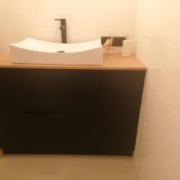 Biała, asymetryczna umywalka nablatowa z czarną armaturą na szafce z ciemnego drewna, uzupełniona o dyfuzor zapachowy i dozownik, na tle białych płytek.