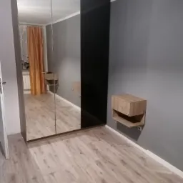 Duża szafa z lustrzanymi drzwiami przesuwnymi i czarnym panelem bocznym, zamontowana w pokoju z szarą ścianą i drewnopodobną podłogą, obok szafka nocna z otwartą półką.