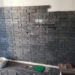 AKR Remonty - Ściana wyłożona szarą imitacją cegły, widoczne gniazdko elektryczne i resztki zaprawy na podłodze, prace w toku.