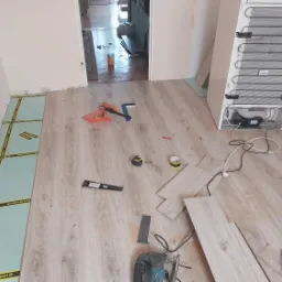 Proces układania paneli podłogowych w pomieszczeniu z widoczną warstwą izolacyjną Mediaexpert, narzędziami, częściowo ułożonymi panelami i stojącą lodówką.