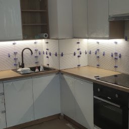 AKR Remonty - Białe płytki ułożone na ścianie nad blatem kuchennym, z użyciem niebieskich krzyżyków dystansowych, podświetlone listwą LED, z widocznym zlewem i płytą indukcyjną.