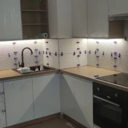 Białe płytki ułożone na ścianie nad blatem kuchennym, z użyciem niebieskich krzyżyków dystansowych, podświetlone listwą LED, z widocznym zlewem i płytą indukcyjną.