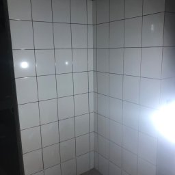 AKR Remonty - Świeżo położone białe, kwadratowe płytki w rogu pomieszczenia, widoczne szare fugi i niedokończone wykończenie przy suficie.