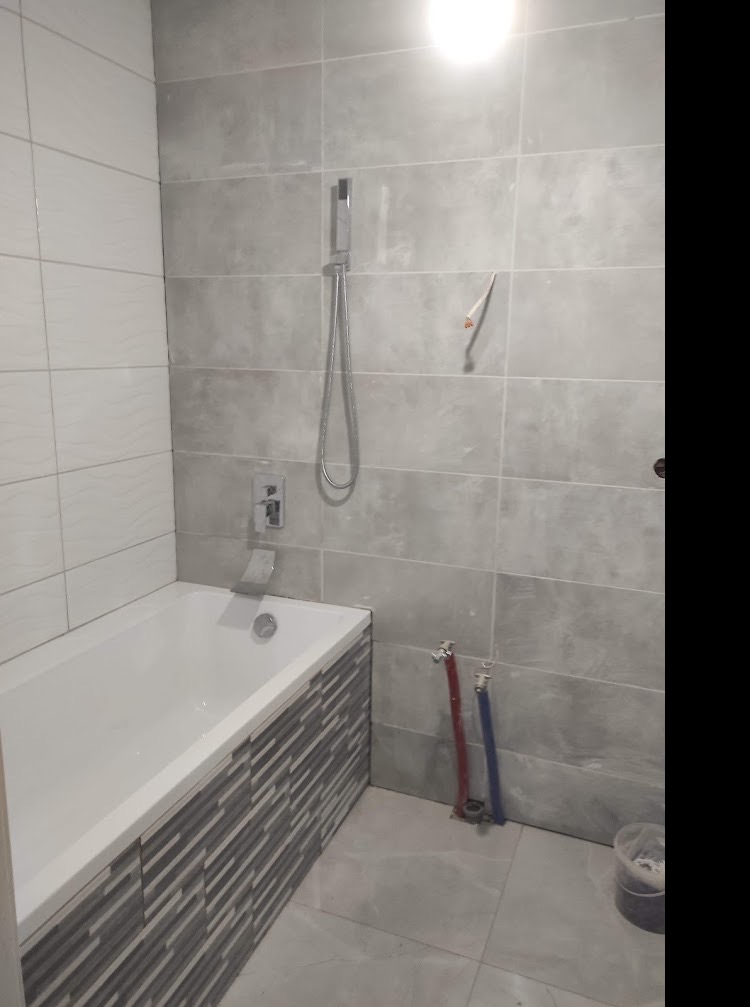 Wykończona łazienka z wanną obudowaną mozaiką, szare płytki na ścianach, widoczne rury instalacji wodnej przed zabudową.